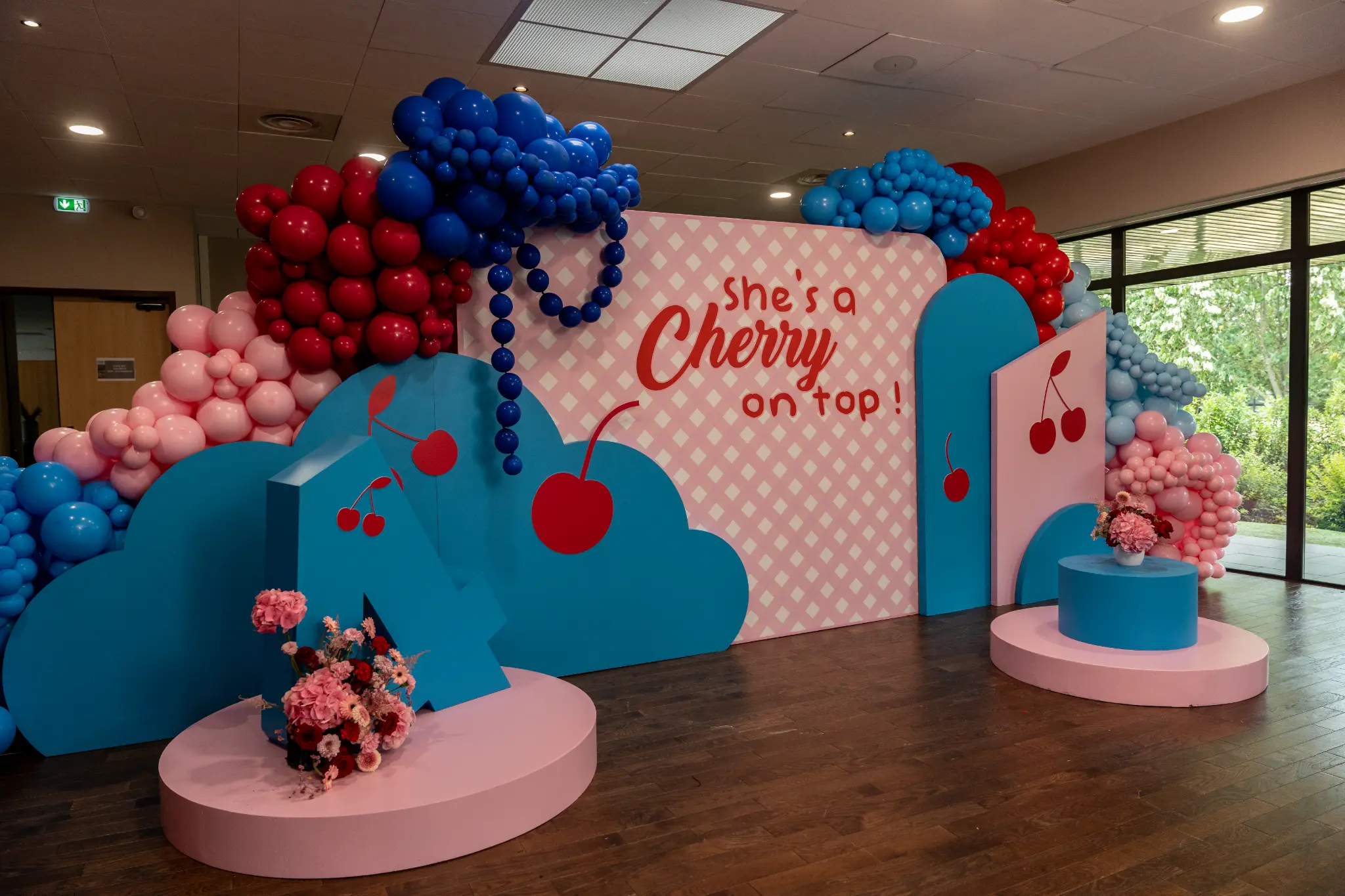 Décor événementiel coloré avec arche de ballons bleus, rouges et roses, panneaux en forme de cerises et fleurs